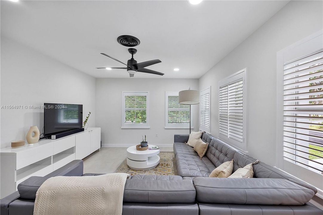 The spacious living room, perfect for watching TV and socializing with friends, features large windows adorned with white plantation shutters, creating a bright and welcoming atmosphere.