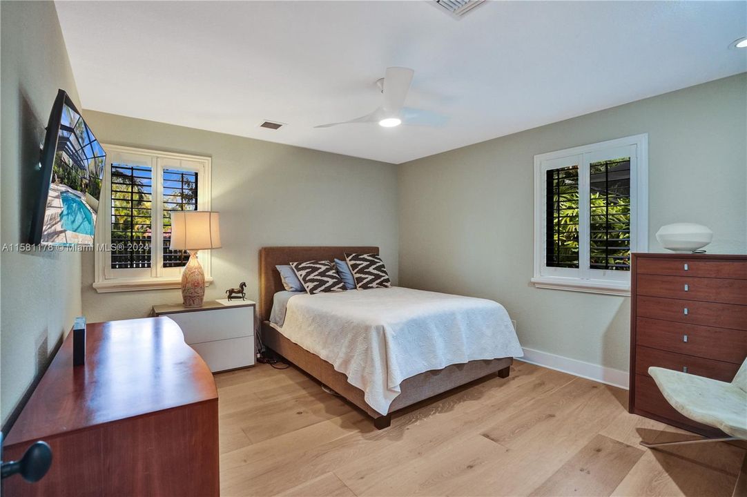Step into a guest bedroom that exudes warmth and tranquility, where natural light floods through expansive windows, casting a soft, inviting glow upon the rich wood floors below. The room feels airy and spacious, with its neutral tones and minimalist decor creating a serene ambiance
