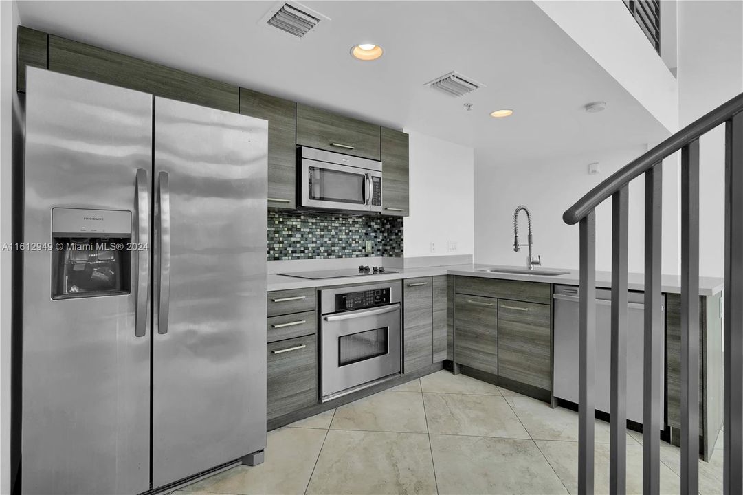 Modern kitchen within a double-height ceiling loft, featuring stainless steel appliances, stylish cabinetry, and a spiral staircase leading to the mezzanine. Bright and airy space.