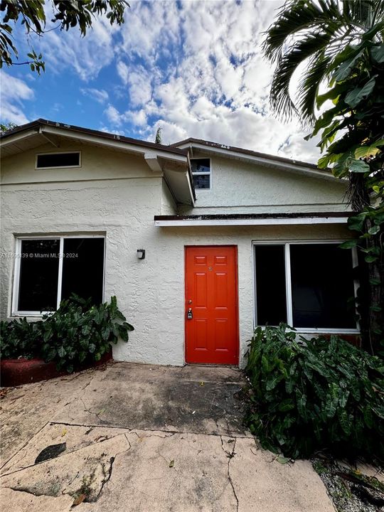 The front of Unit D, a two-story cottage with a Mediterranean-style design, features a distinctive orange door framed by two large rectangular windows, white exterior walls, and a slightly pitched tiled roof with a small window just below the roofline. A concrete pathway leads to the entrance, bordered by lush green plants and shrubs, and palm trees enhance the tropical charm of this quaint and cozy cottage.