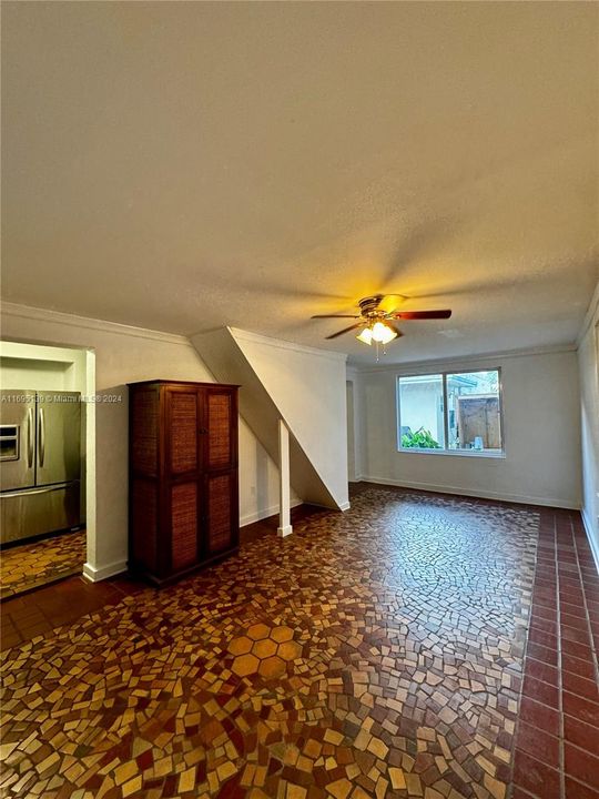 The living room features mosaic-style ceramic tile flooring in brown and beige tones, white walls with a large back window letting in natural light, a ceiling fan with lights, a slanted ceiling for a unique touch, and an open doorway to the kitchen, creating a clean and inviting blend of modern and rustic elements.