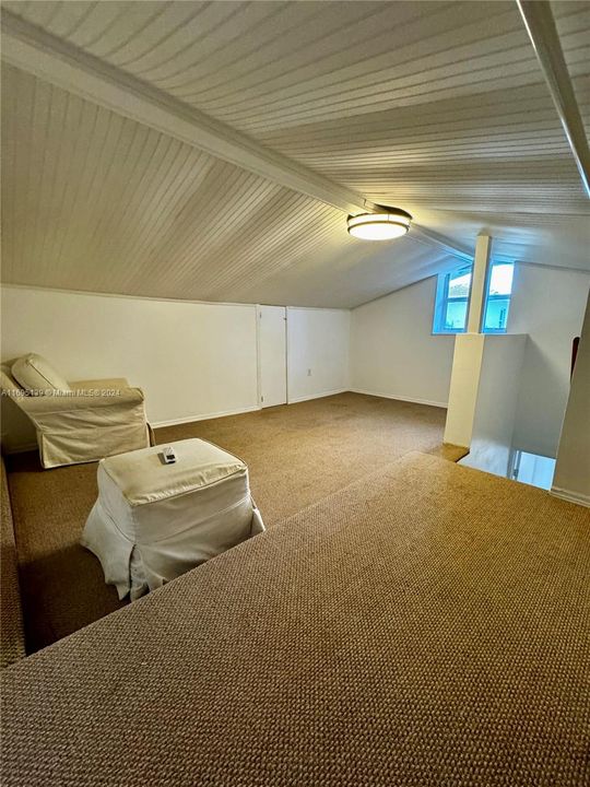 The master bedroom, located on the second floor of the cottage and approximately 250 square feet, features a slanted ceiling with white paneling, light brown carpet flooring, white walls, a round ceiling light, a wall-mounted air conditioning unit, a window providing natural light, and a raised platform for storage, creating a cozy and functional attic-style space.