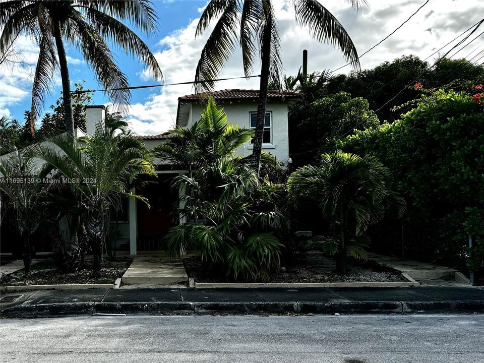 The image shows the entire front structure of the fourplex, which houses Units A, B, and C, while the cottage "Unit D", located in the back, is accessible via the narrow concrete pathway on the right partially obscured by tropical vegetation, including palm trees and lush shrubs.