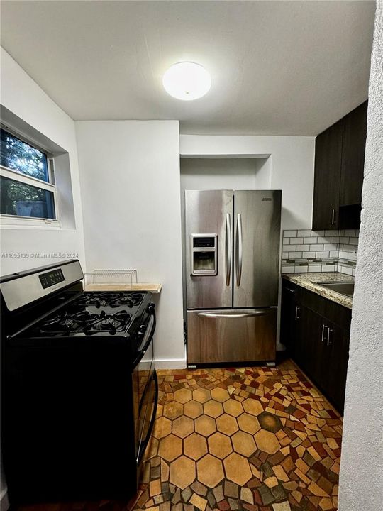 The compact kitchen features hexagonal ceramic tile flooring in warm shades, white subway tile backsplash, dark cabinets, light-colored countertops, stainless steel appliances, a black gas stove under a window, and clean white walls with modern lighting, efficiently utilizing its space.