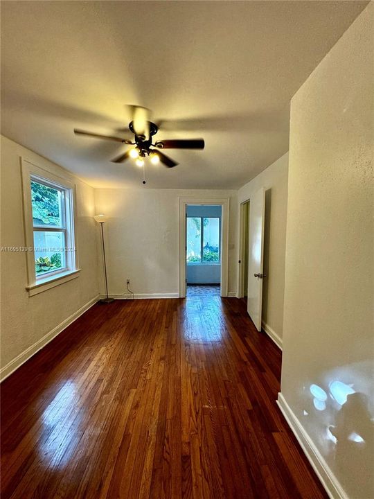 The ground-level bedroom features polished hardwood flooring, white walls and a ceiling fan with a light fixture, two windows filling the space with natural light, and a wall-mounted air conditioning unit. With two doors providing access to the corridor and bathroom, along with a small closet, the room has a clean, minimalistic, and inviting atmosphere.