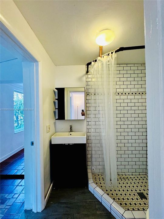 The bathroom, accessible through two separate doors from the ground-level bedroom and the corridor, features a corner shower with white subway tiles and mosaic accents, a sleek black vanity with a square sink and mirror, white-painted walls, and dark flooring, creating a compact, modern, and functional design.