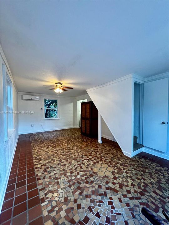 The living room features mosaic-style ceramic tile flooring in brown and beige tones, white walls with a large back window letting in natural light, a ceiling fan with lights, a slanted ceiling for a unique touch, and an open doorway to the kitchen, creating a clean and inviting blend of modern and rustic elements.