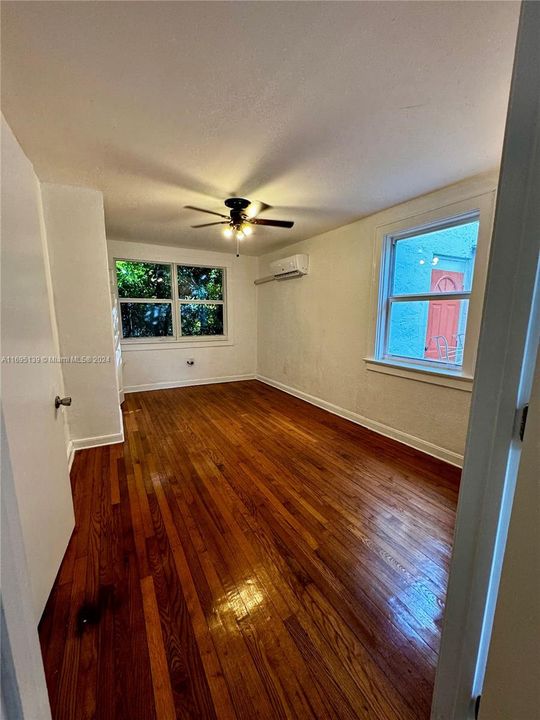 The ground-level bedroom features polished hardwood flooring, white walls and a ceiling fan with a light fixture, two windows filling the space with natural light, and a wall-mounted air conditioning unit. With two doors providing access to the corridor and bathroom, along with a small closet, the room has a clean, minimalistic, and inviting atmosphere.