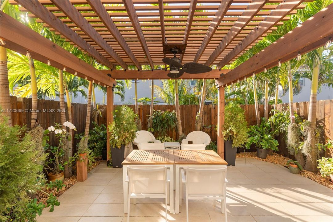 Step into your private tropical oasis! This backyard retreat features a stunning pergola adorned with string lights, perfect for outdoor dining and relaxation. Surrounded by lush landscaping, including towering palms and vibrant greenery, this serene space offers the ultimate blend of privacy and tranquility. Ideal for entertaining or unwinding, the tiled patio and cozy seating area make this a true paradise in your own backyard.