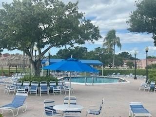 Resort-style community pool area featuring a large blue umbrella, lounge seating, mature landscaping with oak and palm trees, and manicured grounds