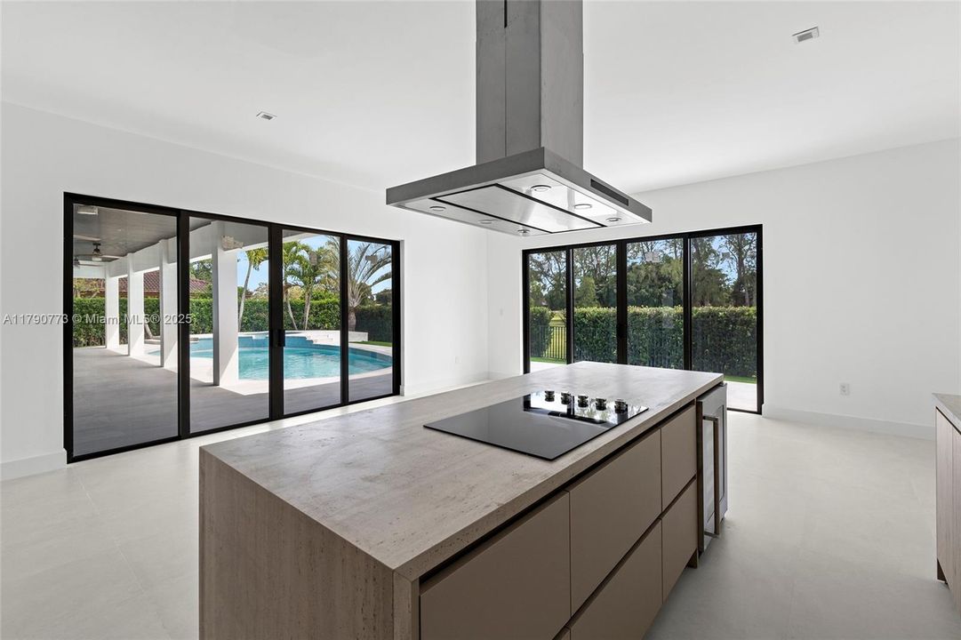 The kitchen offers clean, contemporary design with an electric cooktop, sleek range hood, quartz island with built-in wine cooler, and sweeping views of the pool and private backyard oasis.