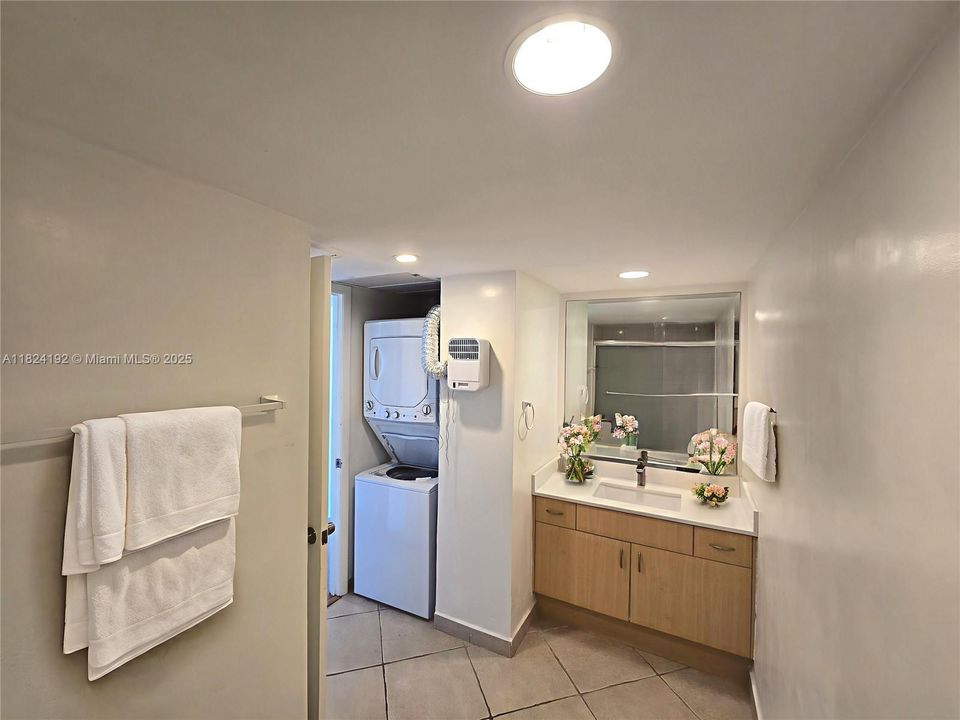 Spacious full bathroom featuring a tub and shower combo with sliding glass enclosure, a large vanity with ample counter space, and an in-unit washer and dryer for added convenience. Bright lighting and neutral tile flooring create a clean, functional environment