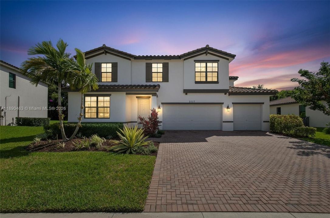 Gorgeous two-story home with a wide paver driveway, three-car garage, and fresh tropical landscaping, all highlighted by warm exterior lighting against a sunset sky — true curb appeal.