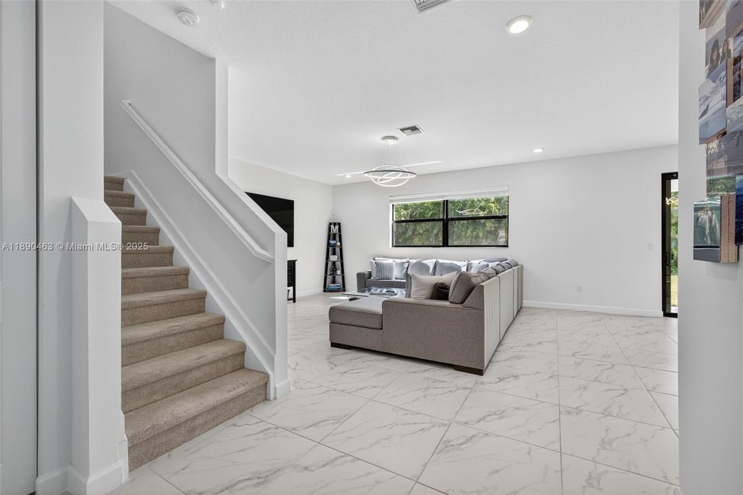 Bright entry atrium with sleek marble-look tile floors that flow into the living space, modern lighting, and an open staircase leading upstairs — a clean, welcoming first impression.