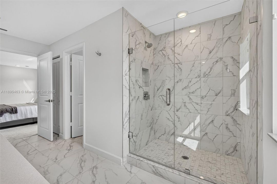 Primary bathroom with sleek marble-look tile floors and walls, featuring a large walk-in shower with frameless glass doors and built-in niche. A window brings in natural light, giving the space a bright, fresh spa feel.