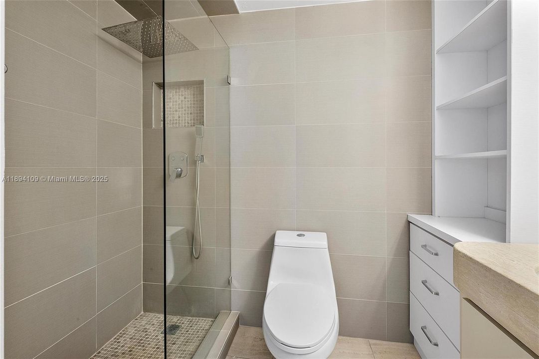 Renovated bathroom has shows ceiling mounted rain shower head, elegant one piece toilet, tile detailed niche.