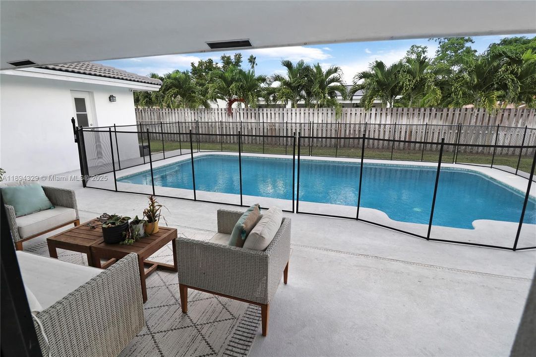 From the kitchen, enjoy a clear and inviting view of the spacious outdoor patio, where a cozy seating arrangement with two chairs and a small table offers a perfect spot to relax. Just beyond, a large swimming pool—safely enclosed by a protective fence—adds a refreshing focal point to the backyard, creating a serene, tropical atmosphere. This picturesque scene brings the outdoors in, enhancing the home's open and airy feel.