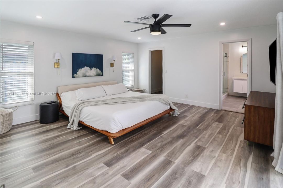 The spacious primary bedroom offers a serene retreat. Wide plank wood flooring in rich, varied tones adds warmth and character, while a ceiling fan with sleek black blades provides comfort and style. To the right, a private en-suite bathroom is conveniently accessible. Opposite the bathroom, a generous walk-in closet provides ample space for wardrobe organization and storage, enhancing the room’s functionality and appeal.