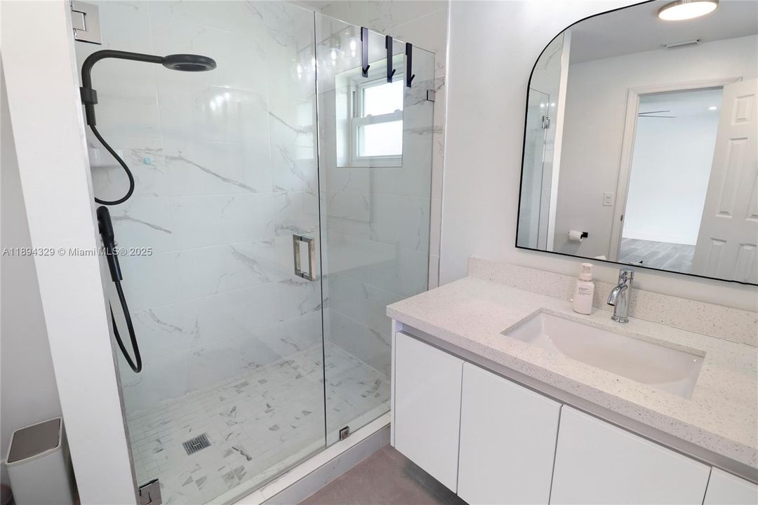 This modern bathroom features a sleek shower-only design, enclosed by clear glass doors with minimalist metal handles. The tiled interior adds a clean, contemporary touch, emphasizing both style and functionality. Opposite the shower, a floating cabinet sink is mounted elegantly on the wall, creating a spacious and uncluttered feel. The white cabinetry and streamlined design complement the overall aesthetic, while an arched mirror framed by wall-mounted lighting adds a sophisticated finish to the space.