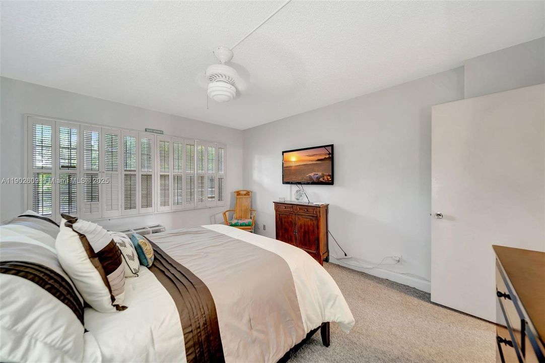 Bright, comfortable bedroom with neutral walls, custom shutters, and soft carpeting. A ceiling fan and generous wall-to-wall windows create a calm, breezy retreat with plenty of natural light