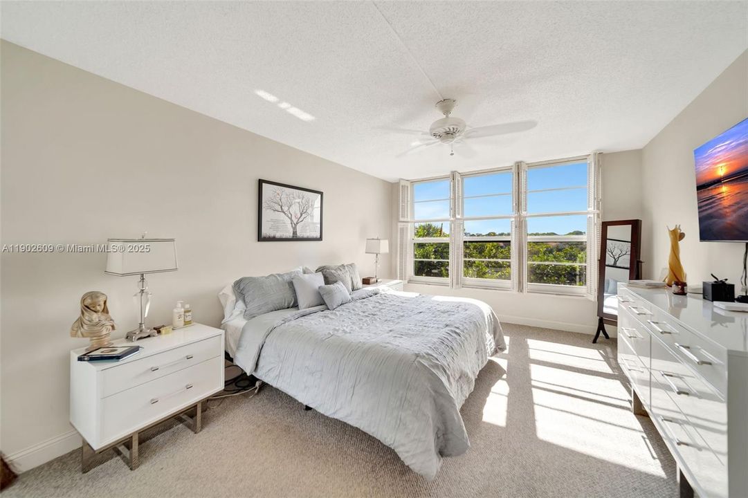 Spacious bedroom with a full wall of windows framing lush green views and incredible natural light. Soft neutral tones and plush carpeting create a calm, comfortable place to unwind.