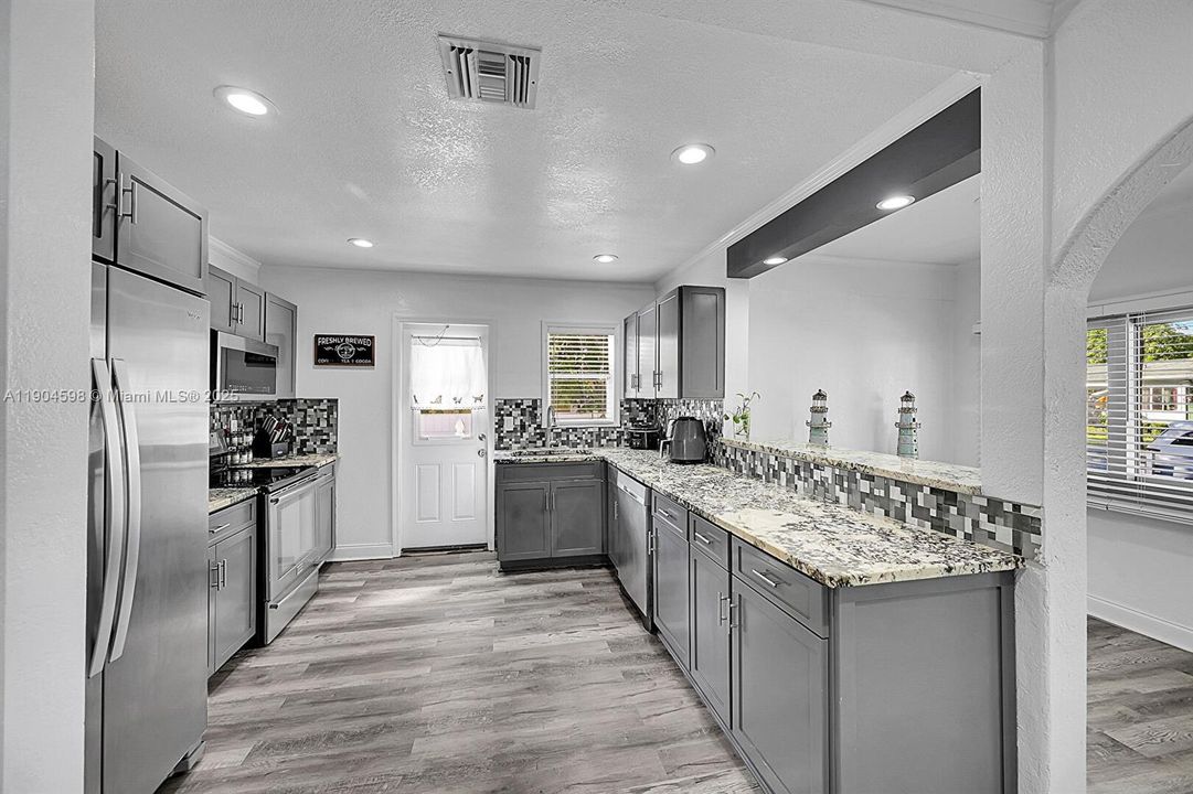 Arched Walkways, Baseboards Wall, Crown Molding, Drywall, Gray Cabinets, Kitchen Peninsula, Natural Light, Opens To Living Area, Pass Through Window, Recessed Lighting, Shaker Cabinets, Stone Countertops, Textured Ceiling, Textured Wall, U Shaped Kitchen, Vent, Wood Finish Floor, Arched Walkways, Drywall, Shaker Cabinets, Textured Ceiling, Vent