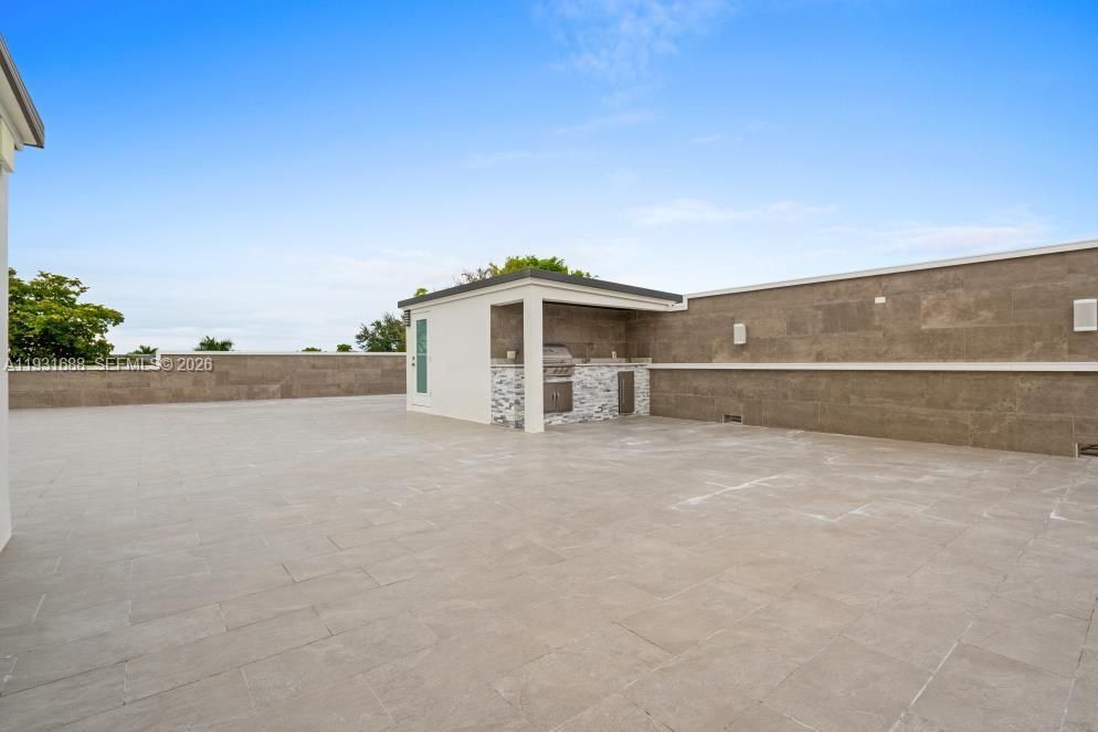 Expansive rooftop terrace featuring large-format tile flooring, perimeter stone wall cladding, and a covered outdoor kitchen equipped with a built-in grill and decorative stone-tiled base