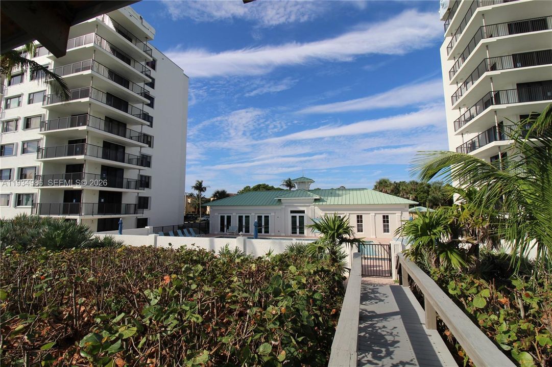 Elevated wooden walkway featuring a metal railing, leading toward a multi-story building complex with a metal-roofed clubhouse and surrounding mature vegetation