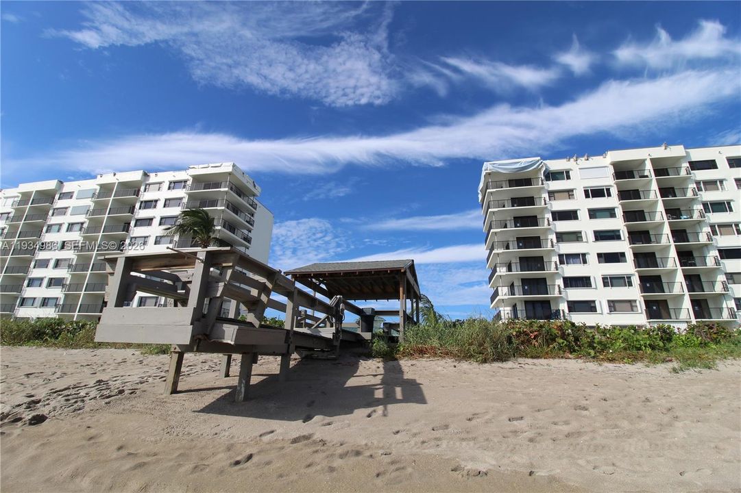 Exterior view of multi-story buildings featuring private balconies and a wooden boardwalk with a covered gazebo structure situated on sandy beach terrain