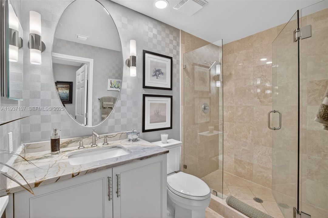 Guest bathroom w/ designer wall coverings, sconces, wall hung mirror. Custom wood vanity. Marble shower walls and vanity counter, Kohler toilet and frameless shower doors