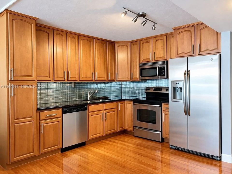 Kitchen featuring wood-finish cabinetry, dark solid-surface countertops, a glass-tile backsplash, stainless steel appliances, and wood-finish flooring