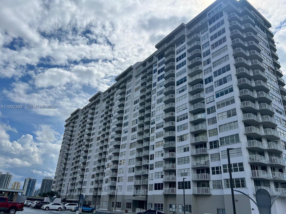 High-rise building exterior featuring a white concrete facade, uniform rows of private balconies with railings, and expansive glass window configurations