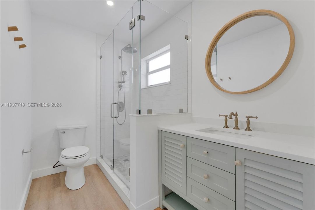 A elegant bathroom featuring a frameless glass walk-in shower with rain head, a sage green louvered vanity with white countertop and brushed gold faucet, a round wood-framed mirror, and light hardwood flooring.