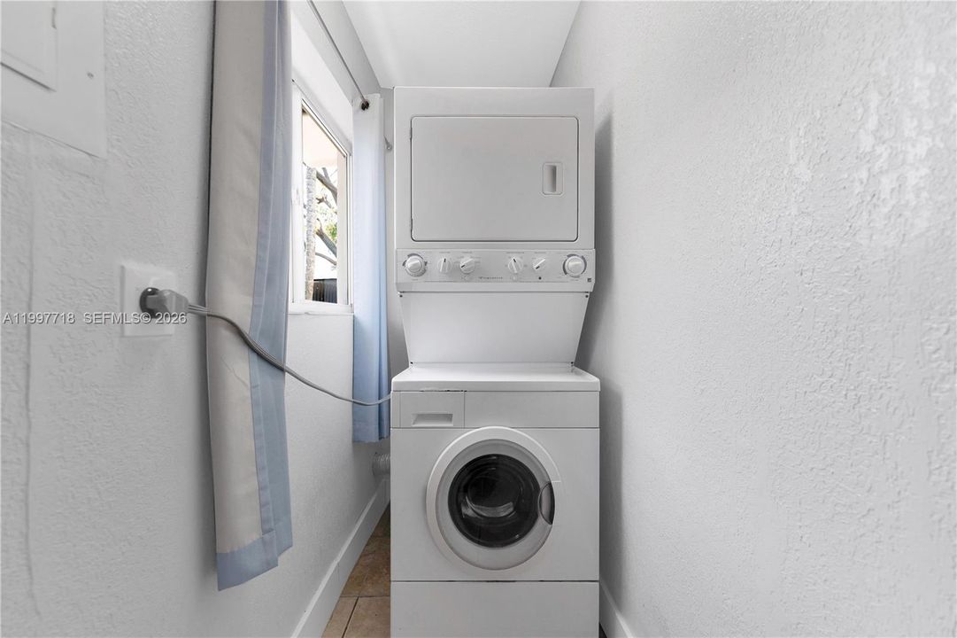 Laundry AreaA narrow laundry nook with a stacked washer and dryer unit tucked against the wall, tile flooring, and a small window with a light blue curtain.