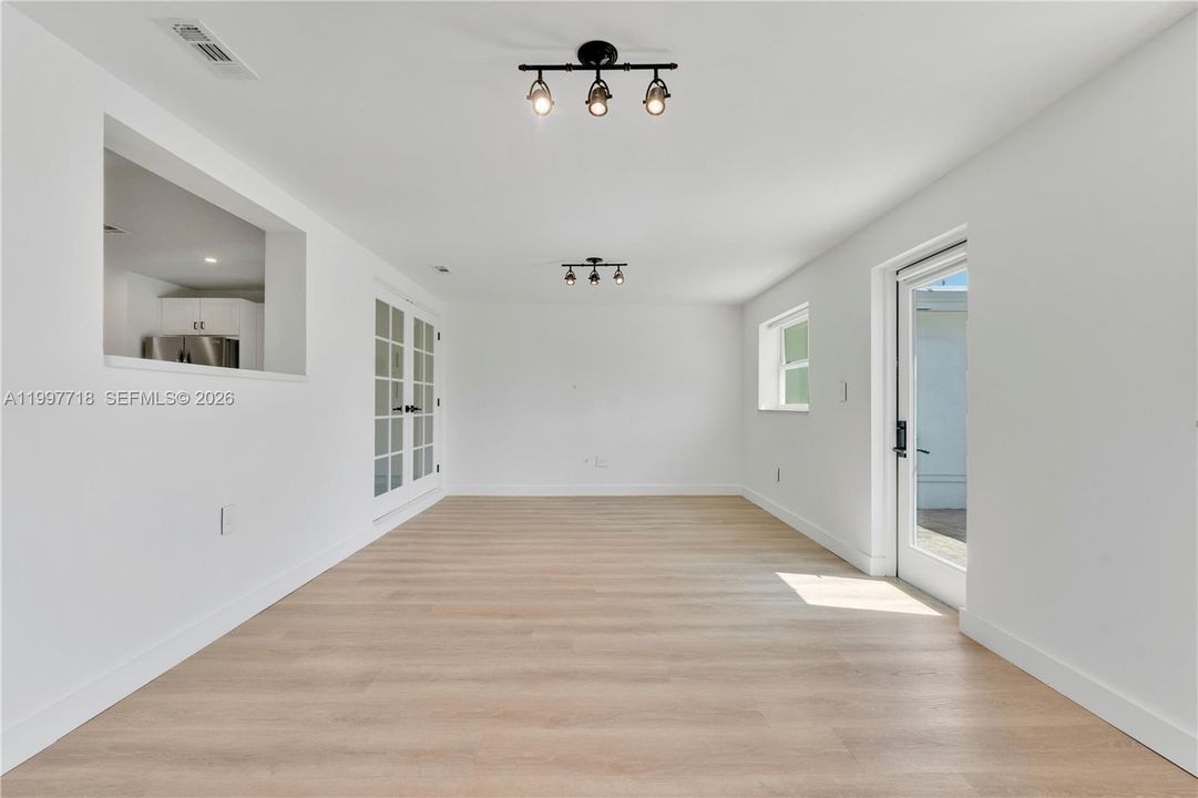 A long, open living space with light hardwood-style flooring, track lighting, and a kitchen pass-through window on the left. Features French glass doors leading to another room, plus a side door opening to the outdoor area.