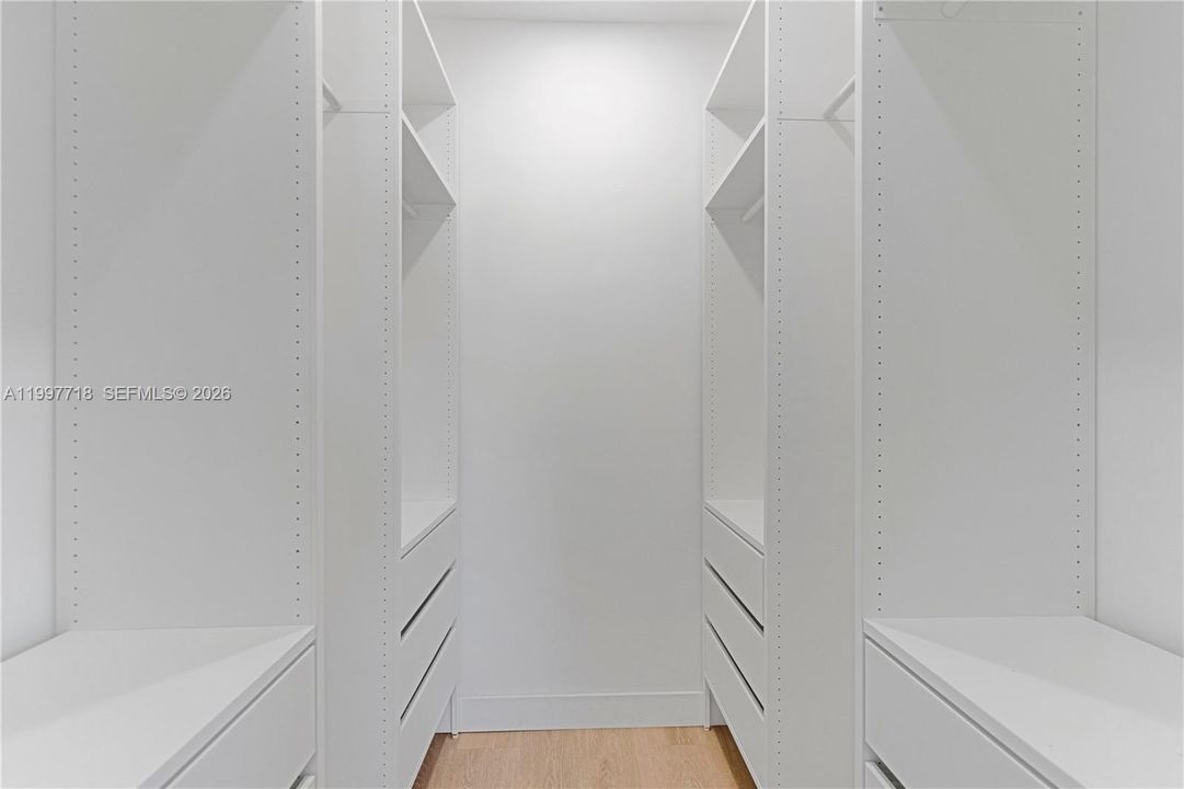 A clean, all-white walk-in closet with built-in shelving, hanging rods, and drawers on both sides, offering ample organized storage space. Light wood flooring runs along the floor.
