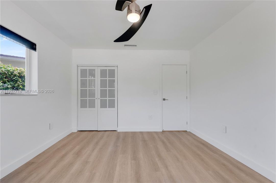 A bright, empty bedroom with light hardwood-style flooring, white walls, a modern two-blade ceiling fan, and two windows with roller shades offering views of the neighborhood outside.