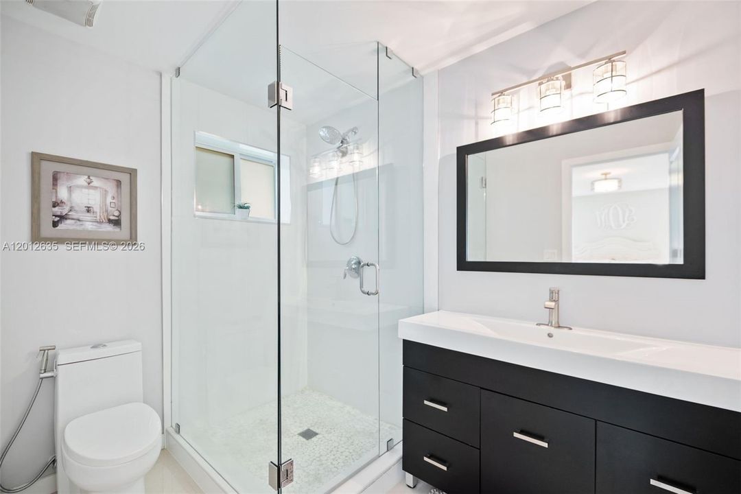 Updated primary en-suite bath with frameless glass-enclosed walk-in shower, pebble tile shower floor, rain showerhead with handheld, espresso vanity with white integrated sink, and designer vanity lighting.