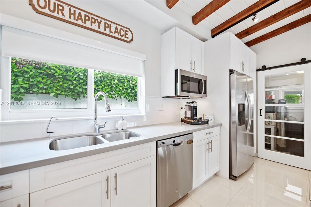 Double stainless steel sink, GE stainless dishwasher and refrigerator, white shaker cabinetry, and garden window overlooking lush landscaping — barn door to utility/laundry room.