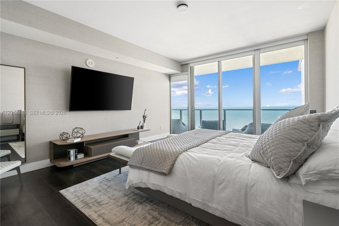 Primary Bedroom – The primary bedroom captures direct water views through floor-to-ceiling glass doors that open to the balcony. A wall-mounted TV, sleek media console, and layered neutral textures create a calm retreat.