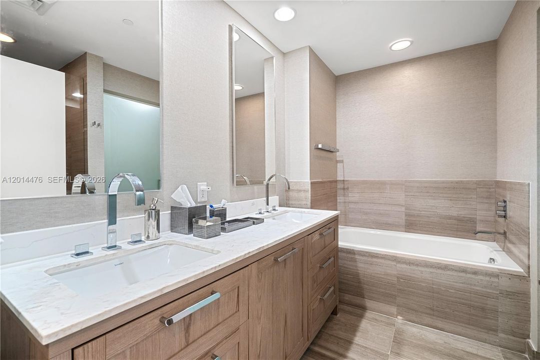 Primary Bathroom – The primary bath features a long wood vanity with a marble-look countertop, dual sinks, and a deep soaking tub framed by warm neutral tile. Large mirrors and recessed lighting create a bright, spa-inspired atmosphere.