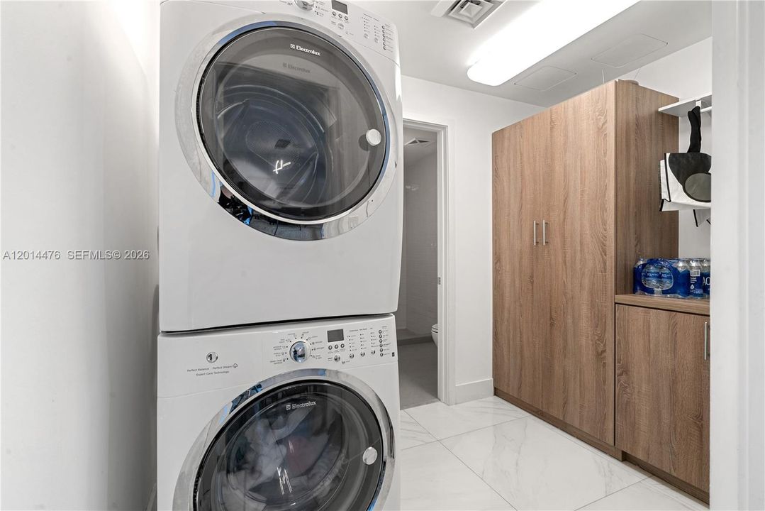 Laundry / Utility Area – A dedicated laundry area features a stacked washer and dryer, glossy marble-look flooring, and built-in wood cabinetry for storage. Bright overhead lighting and a clean layout keep everyday essentials organized and accessible.