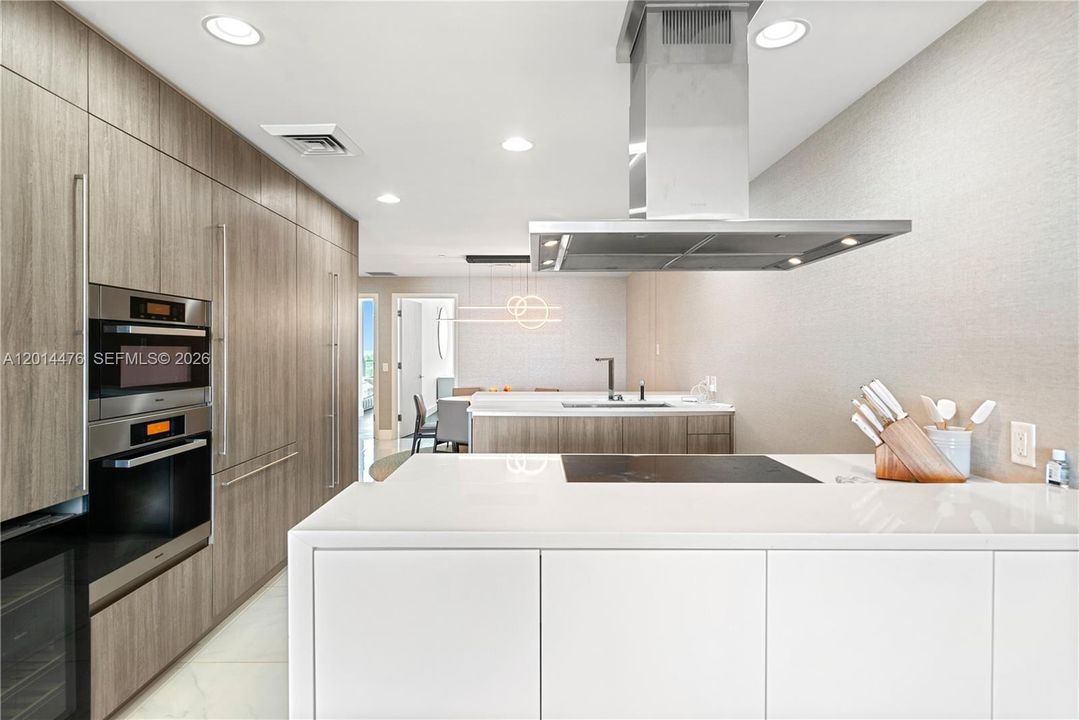 Kitchen – The modern kitchen features white countertops, wood-grain cabinetry, integrated appliances, and a large island with a sleek cooktop and stainless vent hood. Recessed lighting and clean finishes give the space a crisp, contemporary look.