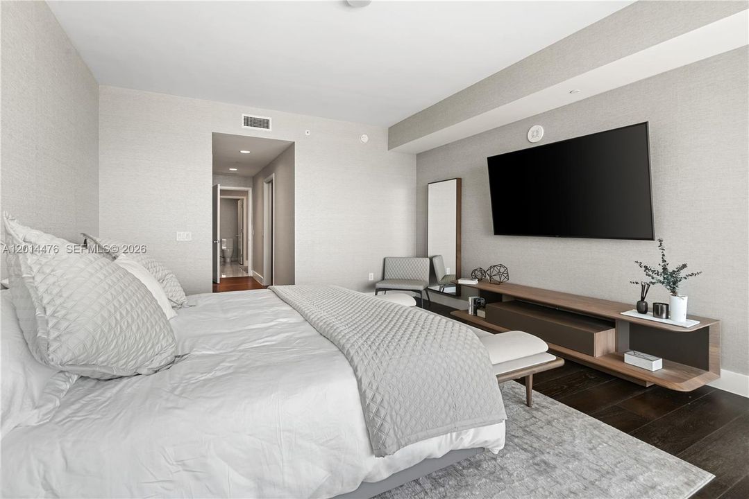 Primary Bedroom – This spacious bedroom features textured wallcovering, dark wood flooring, and a modern entertainment wall. The layout connects easily to the bath and closet areas while maintaining a comfortable, private feel.