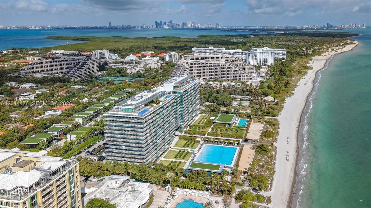 Aerial Waterfront Perspective – This sweeping view shows the building set between the beach and lush residential surroundings, with the skyline visible across the water. The image emphasizes the property’s high-end coastal environment.