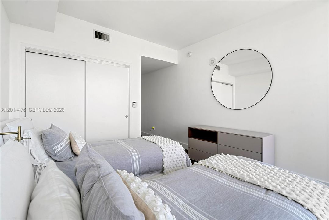 Guest Bedroom – A clean, modern bedroom layout includes a sleek dresser, round wall mirror, and soft neutral bedding. Sliding closet doors and simple finishes keep the space polished and efficient.