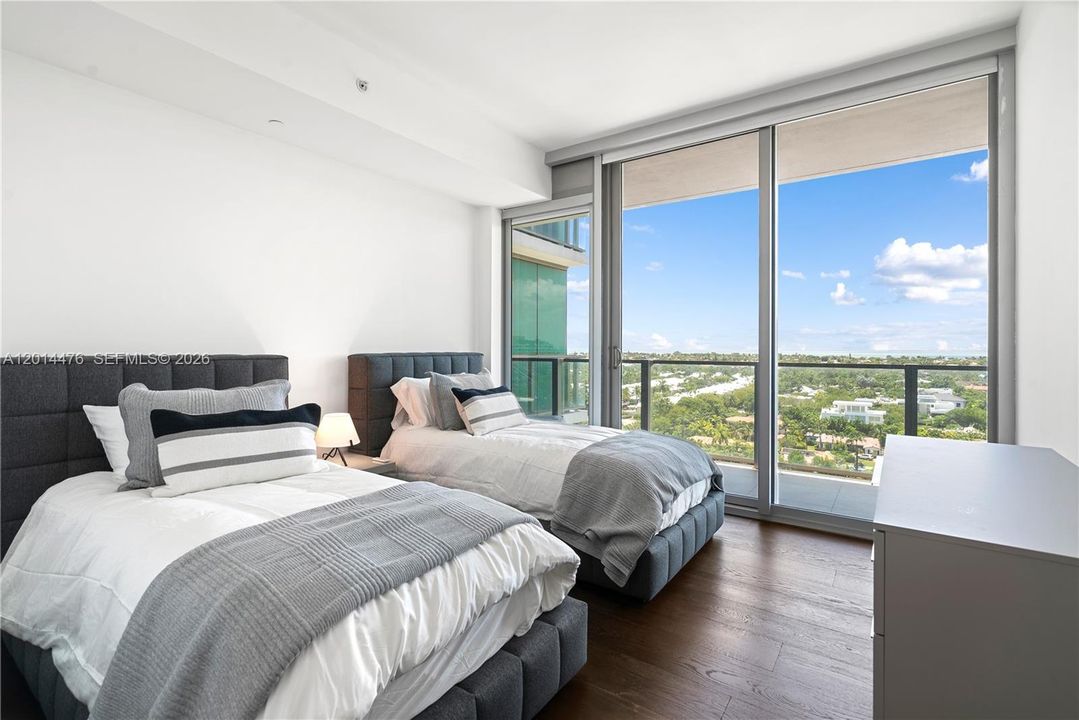 Bedroom with Balcony Access – A second bedroom features floor-to-ceiling glass doors, warm wood flooring, and direct balcony access with sweeping treetop and water views. Soft bedding and neutral tones enhance the calm, coastal feel.