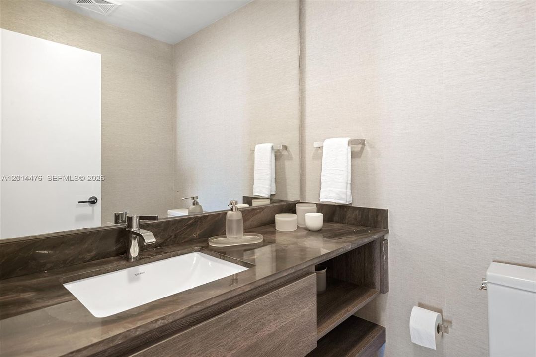 Powder Room – The powder room offers a streamlined vanity with a dark stone countertop, undermount sink, and wall-to-wall mirror. Textured wallcovering and open shelving add both style and function.