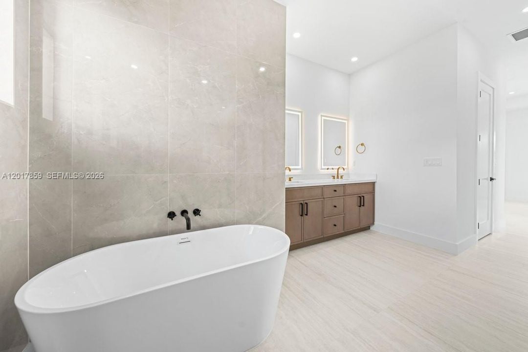 Freestanding soaking tub featuring wall-mounted matte black plumbing fixtures, large-format tile wall surround, double vanity with light-tone wood-finish cabinetry, and recessed lighting