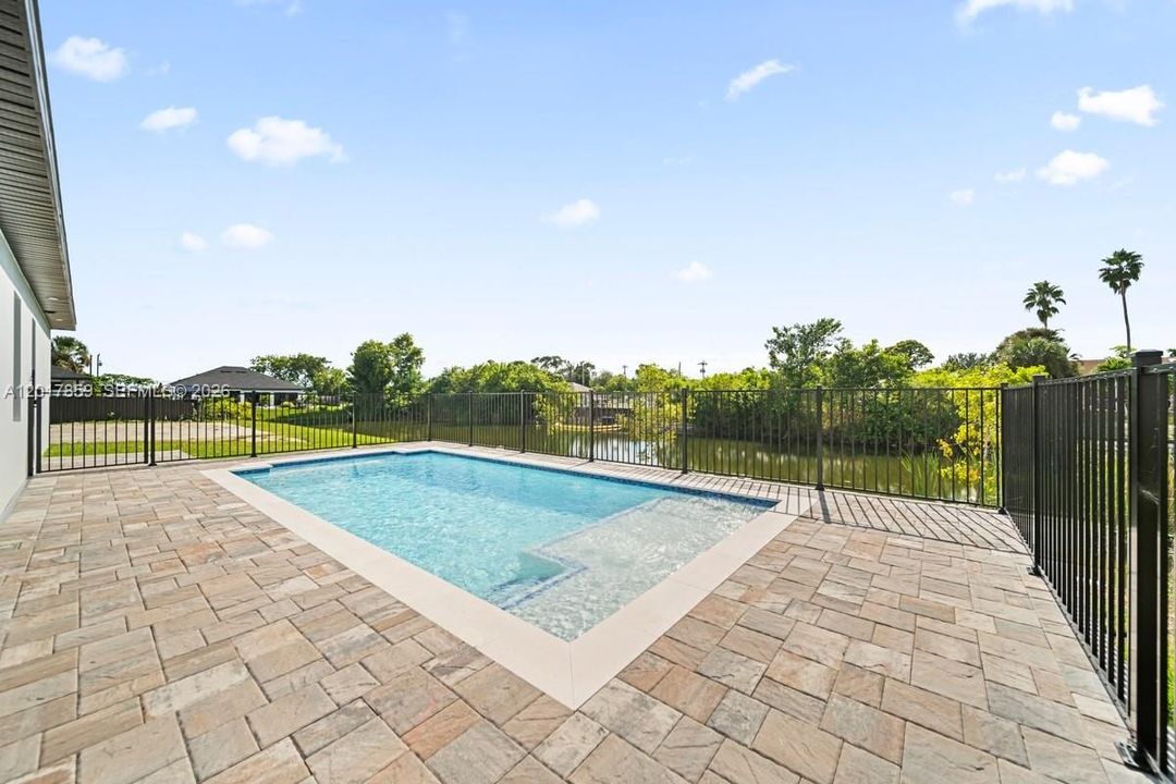 In-ground pool with a tanning shelf, surrounded by a paved patio and black metal perimeter fencing, overlooking a waterfront canal